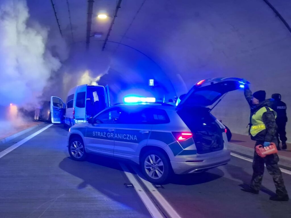 Wnętrze zadymionego tunelu na pierwszym planie radiowóz i policjant